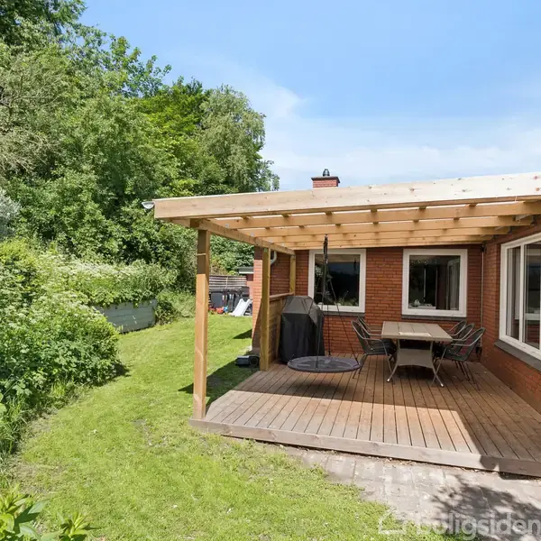 Terrassen har et trædæk med spisebord og stole, beskyttet af et overdække, omgivet af en grøn have og buskads. Rød murstensbygning med store vinduer giver lys.