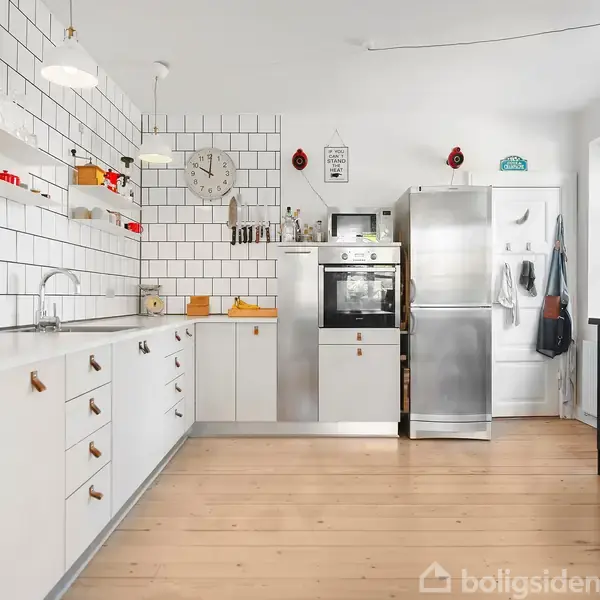 Et moderne køkken med hvide fliser og træskabe, indeholder en indbygget ovn og stålkøleskab. Rummet er lyst med lyse trægulve og vinduer til haven.