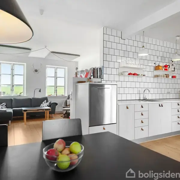 Et moderne køkken-alrum med hvide flisevægge, træbord og grå sofa. På bordet står en skål med frugt. Hylder over køkkenvasken har bøger og køkkenudstyr.