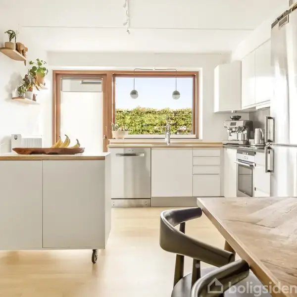 Et lyst, moderne køkken med hvide skabe og træbordplader står i et rummeligt rum. En ø med hjul står centralt, omgivet af store vinduer og grøn udsigt. En træspisebordssæt kompletterer rummet.