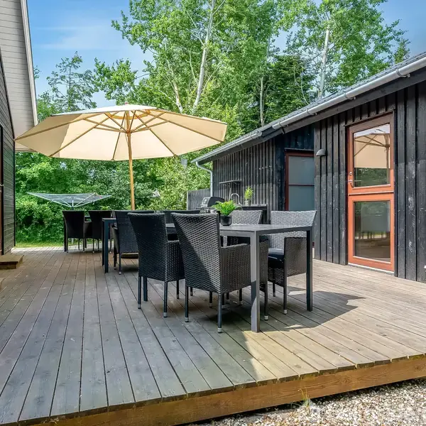 Udendørs terrasse med sorte træmøbler og en stor parasol. Møblerne står på en træplatform ved et sortmalet hus, omgivet af grønne træer i baggrunden.