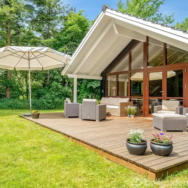 Sommerhus terrasse med siddemøbler under tag; store vinduer bagved. Parasol står på trædæk omgivet af græs og træer, hvilket skaber en naturlig, grøn atmosfære.