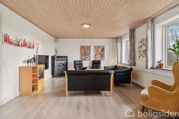 Sort sofaarrangement med lænestole omkring et bord, placeret i en lys stue med træloft og store vinduer. Hylder med dekorationer og billeder hænger på de hvide vægge.