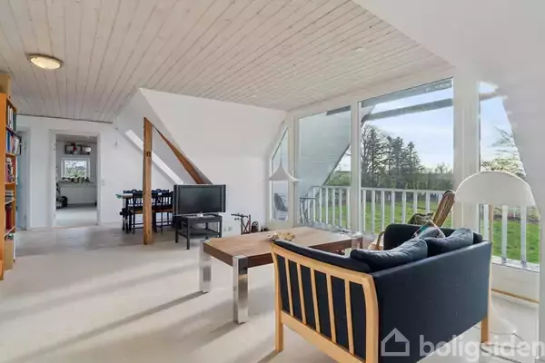 Stue med sofa og bord vender mod store vinduer, der giver udsigt til grønne træer. I baggrunden ses en spiseplads. Rummet er lyst og moderne.