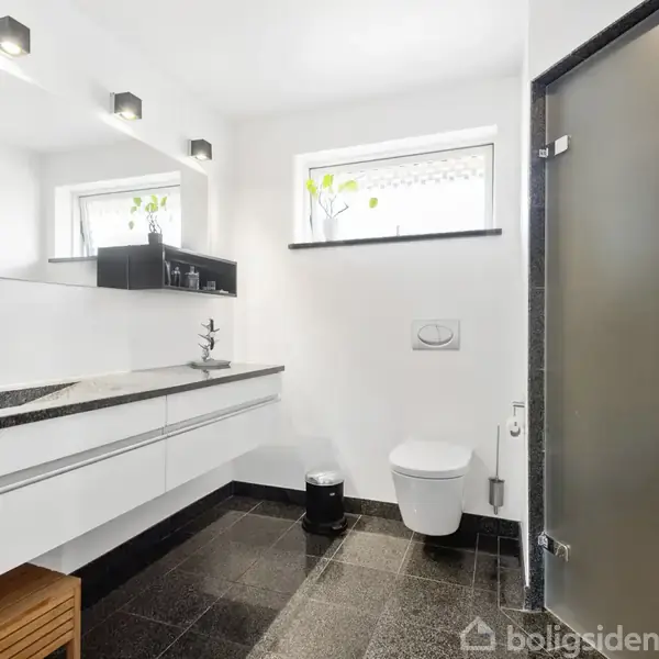 Badeværelse med hvidt interiør, hvor håndvasken er integreret i en sort granitbordplade. Over vasken hænger et spejl, og der er en vægmonteret toilet. Vinduer lukker dagslys ind.