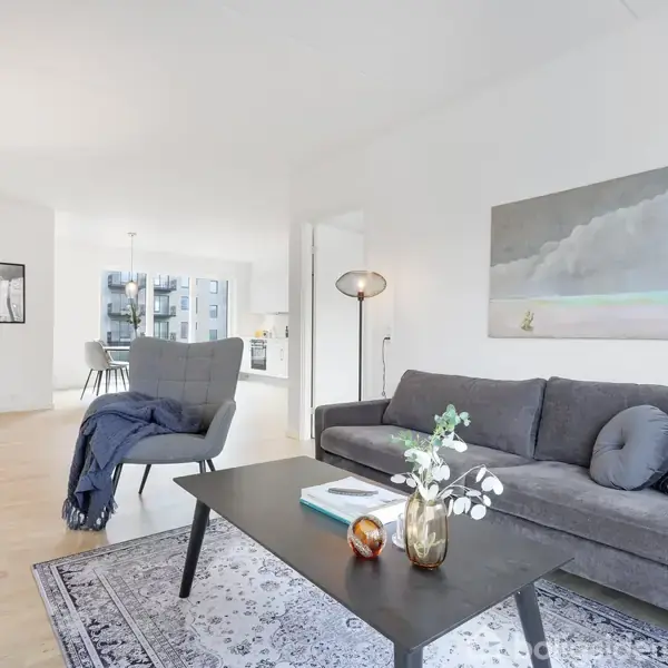 Grå sofa med puder står foran et sort sofabord med dekorationer. Rummet er lyst og rummeligt med moderne indretning. En lænestol og kunst på væggene supplerer set-uppet.