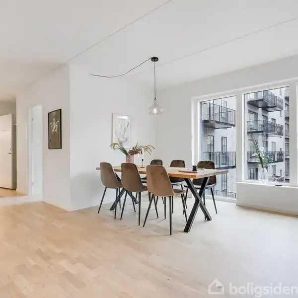 Spisebord med seks stole står i et lyst rum med trægulv. Store vinduer viser udsigten til en moderne bygning med altaner. En enkelt lampe hænger over bordet.