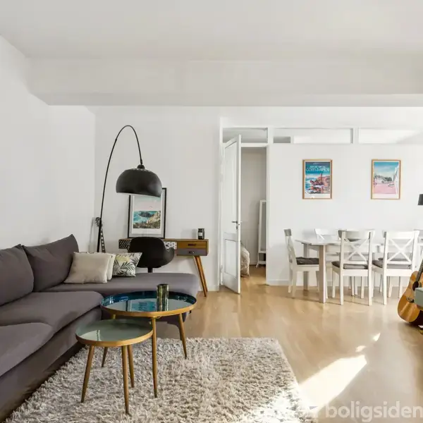 Grå sofa står i stue ved siden af hvilestol og gulvlampe, mens spisebord med hvide stole ses i baggrunden. Trægulv og billeder på væggen udgør konteksten.