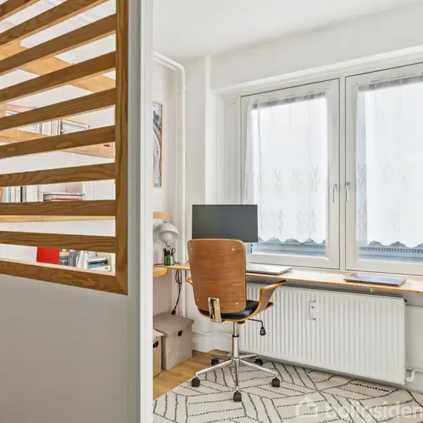 Et skrivebord med en computer og kontorstol står foran et vindue med gardiner. Rummet er lyst og har en tæppebelagt trægulv samt en radiator under vinduet.