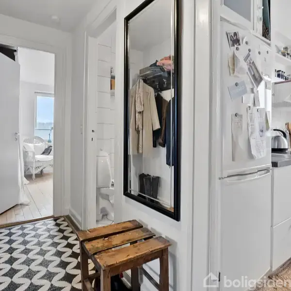 En gang med trægulv og mønstret tæppe, hvor jakker hænger. Der er et spejl, en bænk, og til højre et køkken med køleskab og komfur.