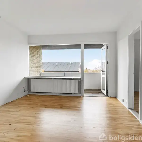 Stue med trægulv, der åbner sig mod en altan gennem en glasdør. Rummet er hvidmalet med store vinduer, der giver udsigt over et tag.