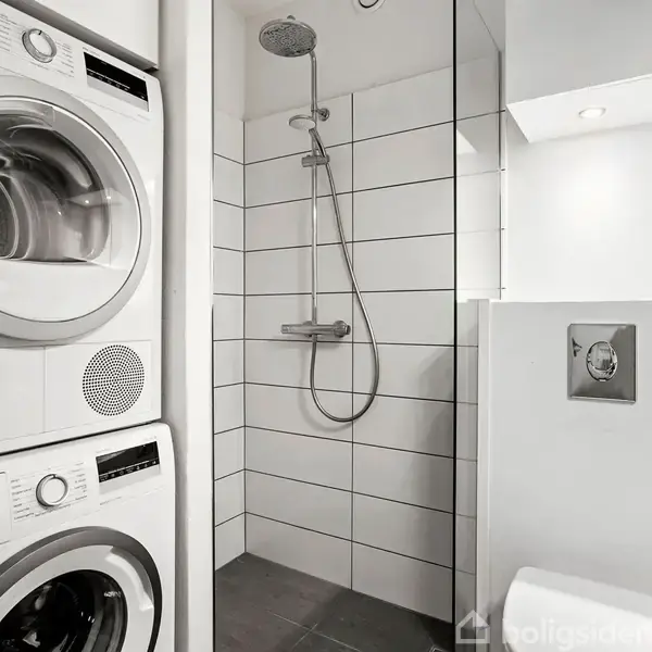 En vaskemaskine og tørretumbler er stablet. Bruseren står til venstre i en bruseniche med hvide fliser. Rummet har grå gulvfliser og en indbygget skylleknap til en toilet.