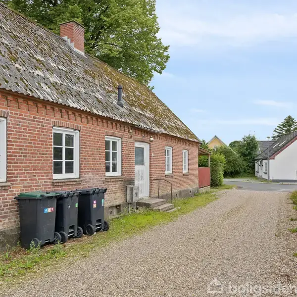 Et rødt murstenshus står langs en grusvej med affaldsbeholdere foran. Flere vinduer og en hvid dør ses på husets facade. Omgivet af grønne buske og en byvej.