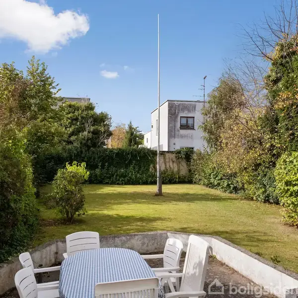 En hvid havestolssæt står udenfor i en terrasseområde. Bagved ses en græsplæne med en flagstang omgivet af frodige buske og træer under en blå himmel med få skyer.