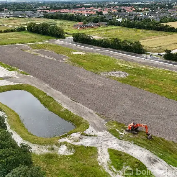 Gravemaskine arbejder på en stor, nyligt forberedt mark med en lille sø i nærheden, omgivet af grønne områder og nogle fjerne bygninger i baggrunden.