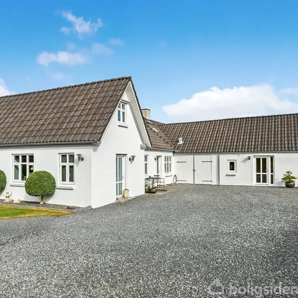 Hvidt hus med gråt tag står stationært på en grusvej, omgivet af græsplæne og blå himmel.