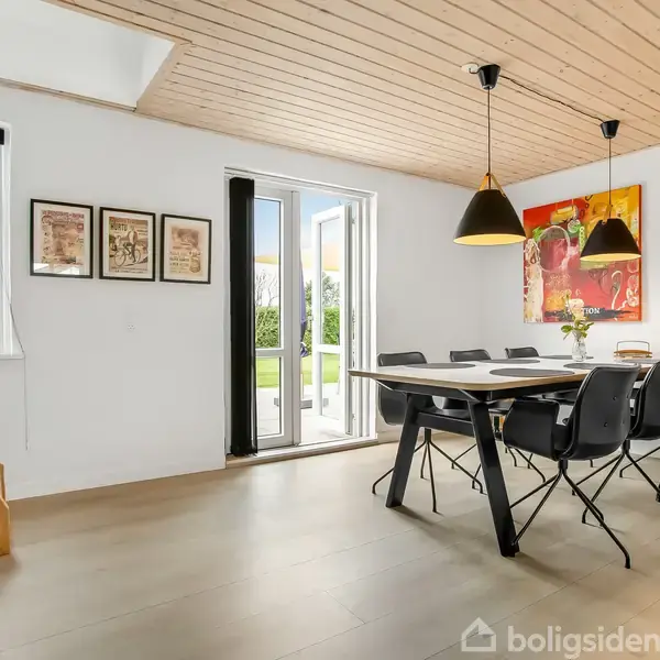 Et spisebord står i en lys spisestue med sorte stole omkring. Store vinduer åbner mod en grøn have. Kunstværker hænger på væggene, og loftslamper giver belysning.