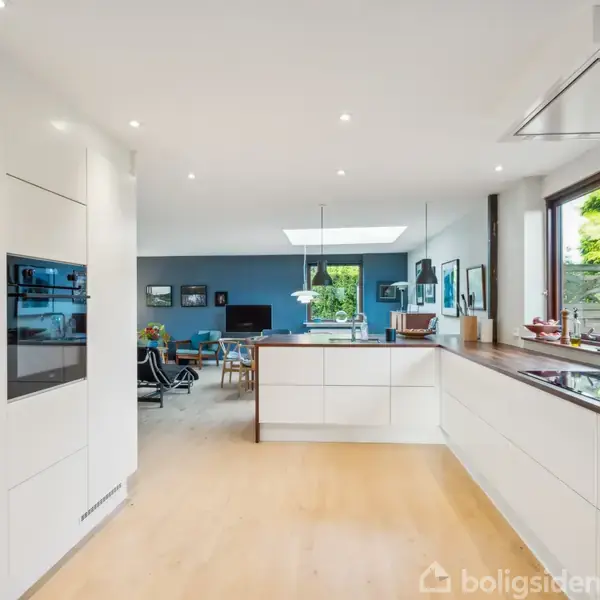 Et moderne køkken med hvide skabe og indbygget ovn. Køkkenet åbner op til en spisestue med blå vægge og grøn udsigt gennem store vinduer.