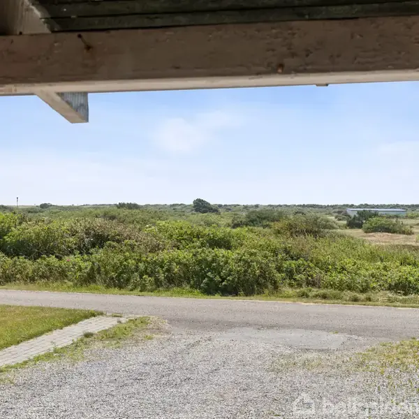 Tagudhuse ligger stille ved en landevej omgivet af grønne buske og en stor, åben himmel i det fjerne. Grusstykker og en asfaltvej forbinder stedet med omgivelserne.