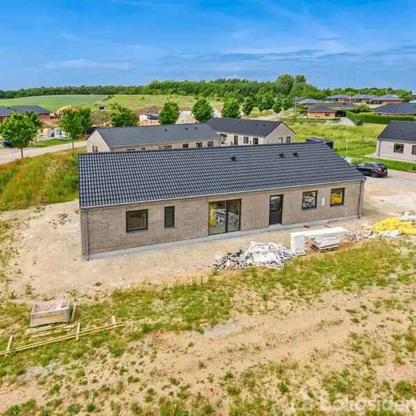 Et nybygget, enetages hus med gråt tag står på en ujævn byggeplads omkranset af græs og grus. Baggrunden viser en forstadsområde med flere lignende huse under blå himmel.