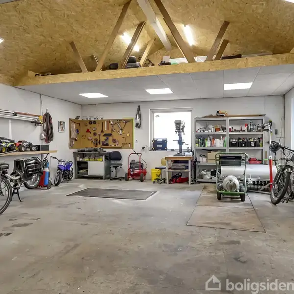 En garage indeholder cykler, værktøj og maskiner. Der er arbejdsborde langs væggene og reoler med forskellige redskaber. Loftet har opbevaringsplads. Det ser organiseret og funktionelt ud.