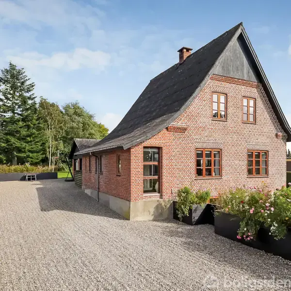 Rødt murstenshus med steept tag står stille ved en grusbelagt indkørsel. Omkringliggende have med blomster og buske; træer og en blå himmel i baggrunden.