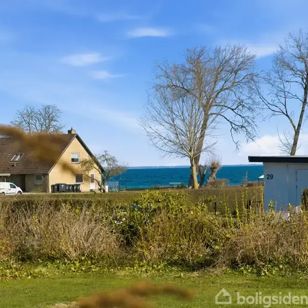 Hus ved havet med et sort tag, omgivet af træer og buske i foråret. Huset er beliggende tæt på vandet, med havet synligt i baggrunden. Nummer 29 ses på et hegn.