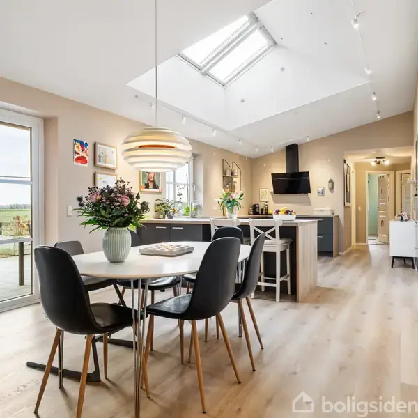 Rundt spisebord med sorte stole står i en moderne, lys køkken-alrum. Ovenlys vindue giver naturligt lys, mens grønne planter pynter rummet. Køkkenø med barstole ses i baggrunden.