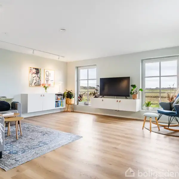 Stue med grå sofa og blå lænestol på trægulv, fjernsyn på hvid reol foran vinduer, vægkunst og planter omkring, skaber en lys og moderne indretning.