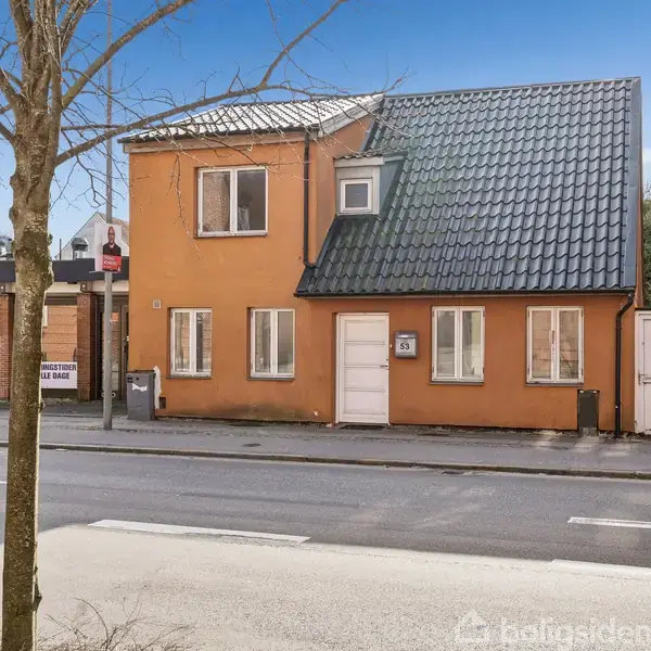 Et orange hus med sort tag står langs en byvej. Udenfor ses en parkeringsskilt og et træ. Til venstre ligger en pizzarestaurant ved navn "Anna Pizza."