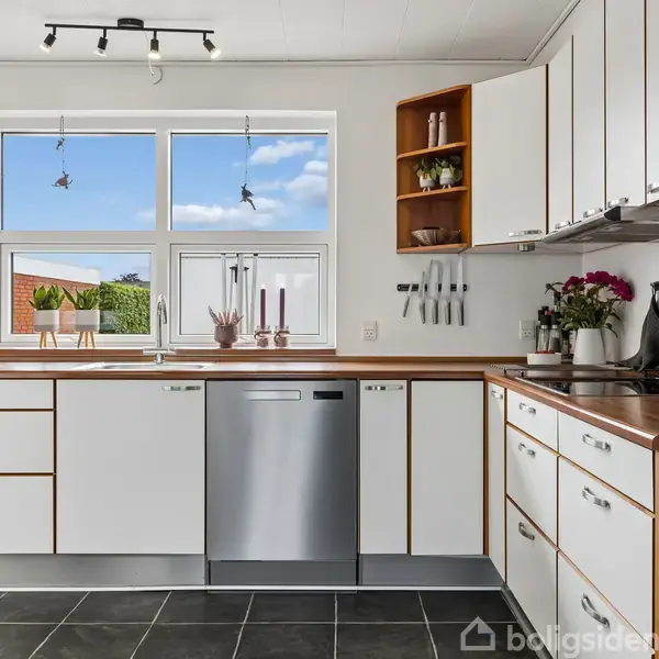 Køkken med hvidt kabinet, træbordplader og mørke fliser. Opvaskemaskine i midten, blomster og køkkenredskaber på bordet. Store vinduer med udsigt til en solrig have.