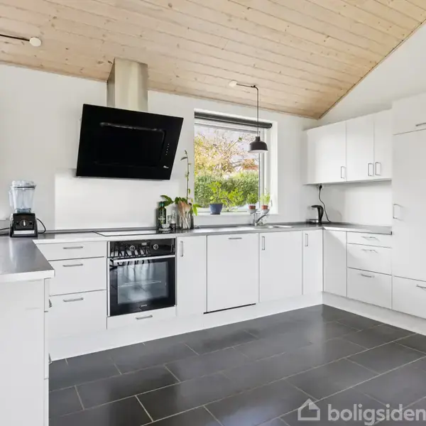 Et moderne, funktionelt køkken med hvide skabe og ståldetaljer. Der er en sort ovn og kogeplade, to vinduer med udsigt til en have, samt sorte gulvfliser og lyse trælofter.
