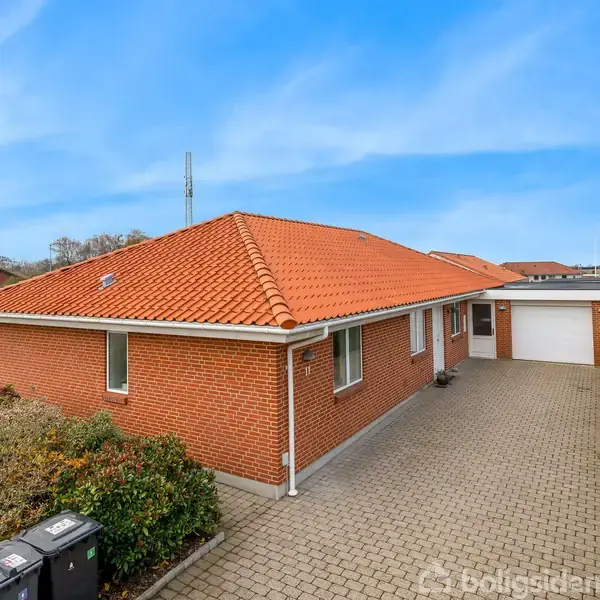 En brunmurstensvilla med rødt tegltag står på en flisebelagt indkørsel, omgivet af velplejet græsplæne og buske, under en klar blå himmel.