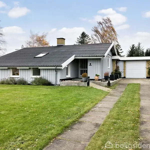En hvid villa med sort tag ligger på en velplejet græsplæne. Huset har en stenbelagt indkørsel, der fører til en garage. Omkring er der træer og buske.