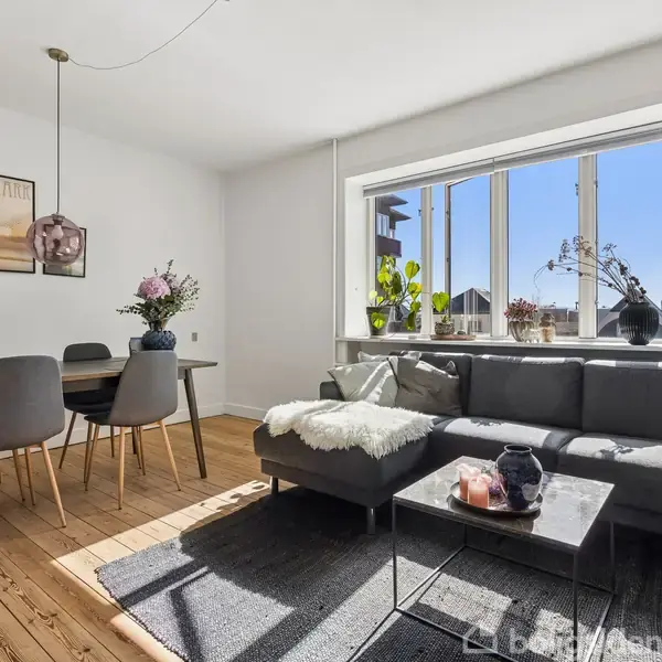 En stue med en grå sofa fyldt med puder, står ved et stort vindue med planter. Ved siden af står et spisebord med fire stole, blomster og kunst på væggen.