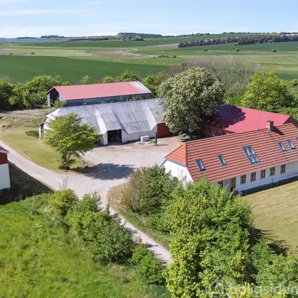 En gård med rødt tag og flere bygninger står omgivet af grønne marker og træer. En grusvej fører til ejendommen, der er omgivet af naturskønne landskaber.