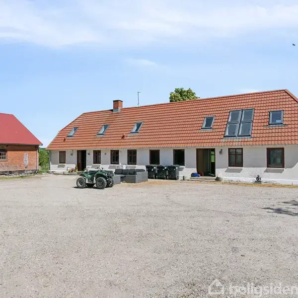 En hvid bygning med rød tag, omkranset af grusplads, står med skraldespande og en trailer foran. Til venstre en anden rødbygget struktur. Klar blå himmel.