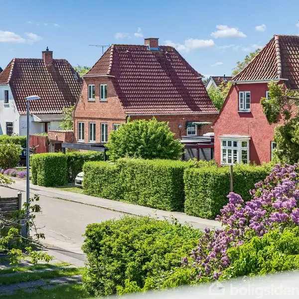 Røde murstenshuse står stille, omkranset af veltrimmede hække og blomstrende buske, i et solrigt boligområde med blå himmel og spredte skyer.