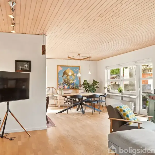 Stue med fladskærms-TV på stativ, der vender mod sofa. I baggrunden et spisebord og stole. Rummet har lyst trægulv og loft med indbygget belysning.