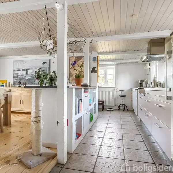 Et køkken i et hjem med hvidt træloft og fliser på gulvet. Køkkenet har hvide skabe, planter på bordet, og en åben hylde. Ved siden af er der en spisestue med trægulv.