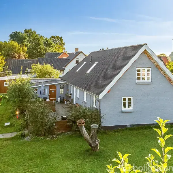 Et gråt hus med skråt tag står i en grøn have. Foran er en flisebelagt terrasse og forskellige buske. Omkring ligger et villakvarter med flere træer i baggrunden.