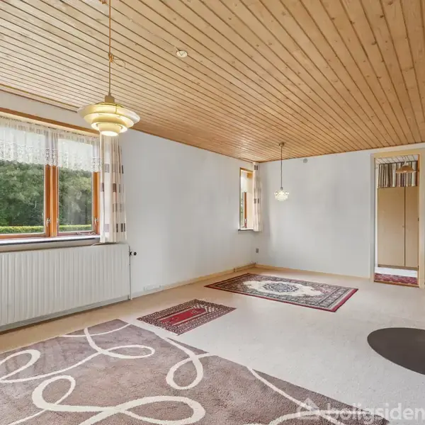 Stue med brændeovn står i hjørnet, under et fyrretræspanel-loft med hængende lamper. Store vinduer med gardiner viser grøn udsigt, og dekorative tæpper dækker trægulvet.