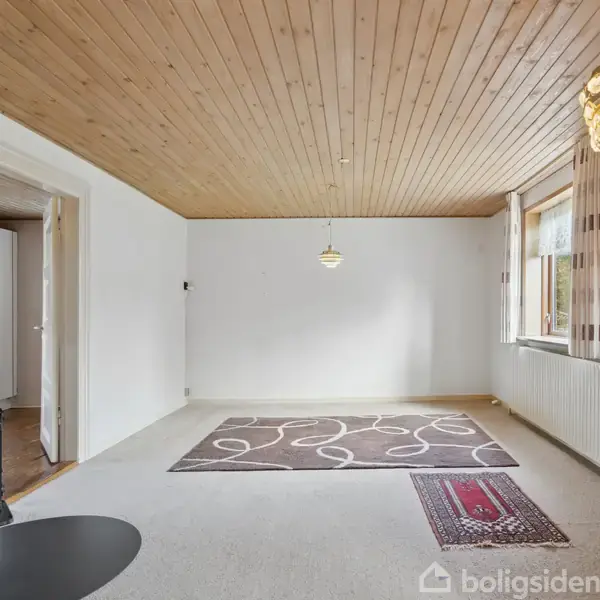 Stue med træloft, to tæpper på gulvet, en sort brændeovn i hjørnet og to lysekroner hængende fra loftet. Vindue til højre med stribede gardiner.