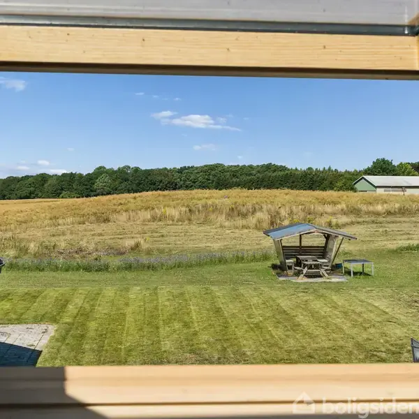 Vindue åbent. Udsigt over nyslået græsplæne med en pavillon og mark i baggrunden. Omgivet af en landejendom og bevoksning, giver en følelse af landlig idyl og natur.
