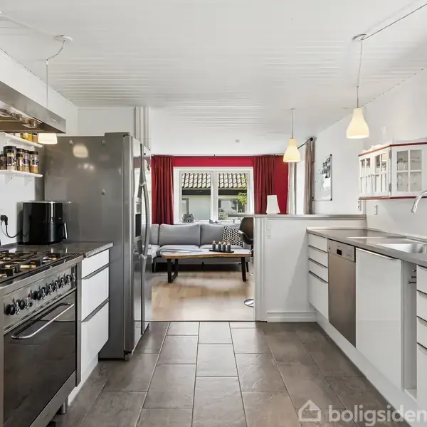 Køkkenet har moderne apparater, herunder et stort gaskomfur med emhætte og køleskab, med åben forbindelse til en stue, der har røde gardiner og lysegrå sofaer.