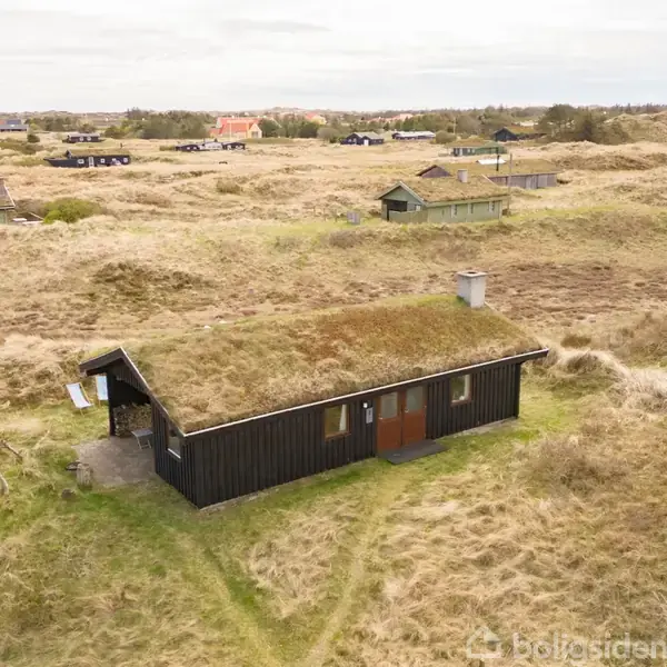 Et sort træhus med græstag ligger i et åbent klitlandskab. Flere små huse ses i baggrunden blandt klitterne.
