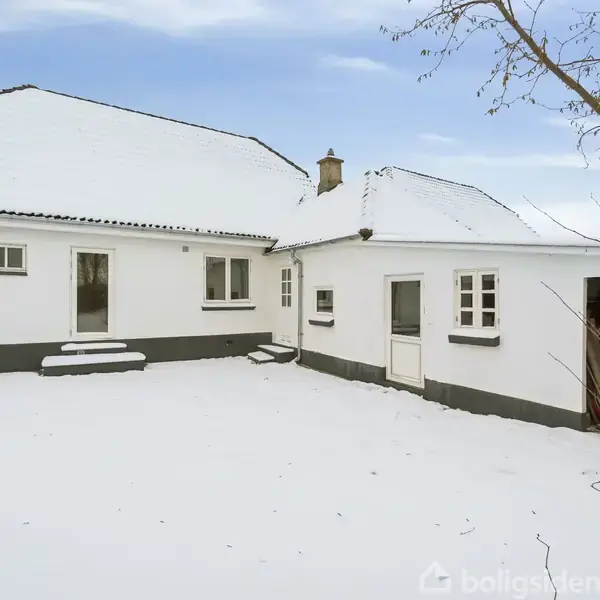 En hvidmalet bygning med skråt tag står i en snedækket gårdsplads. Der er tre vinduer og en åben garage. I baggrunden ses en klar, blå himmel.