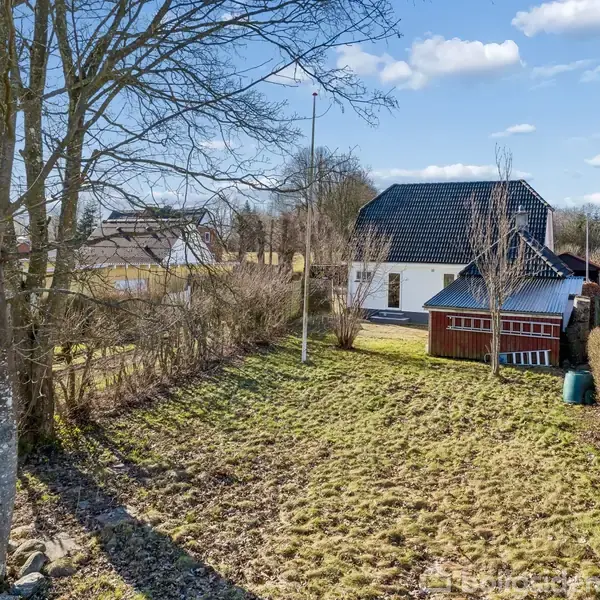 Et hus med sort tag står omgivet af en have med nøgne træer og en bard grønlignende græsplæne. En rød udbygning ses ved siden af huset under en blå himmel med skyer.