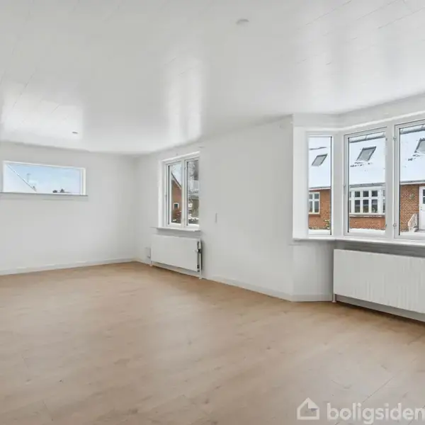 Et lyst, tomt værelse med trægulve. Store vinduer lukker naturligt lys ind, og varmeapparaterne under vinduerne. Udenfor ses rækkehuse med skrå tage dækket af sne.