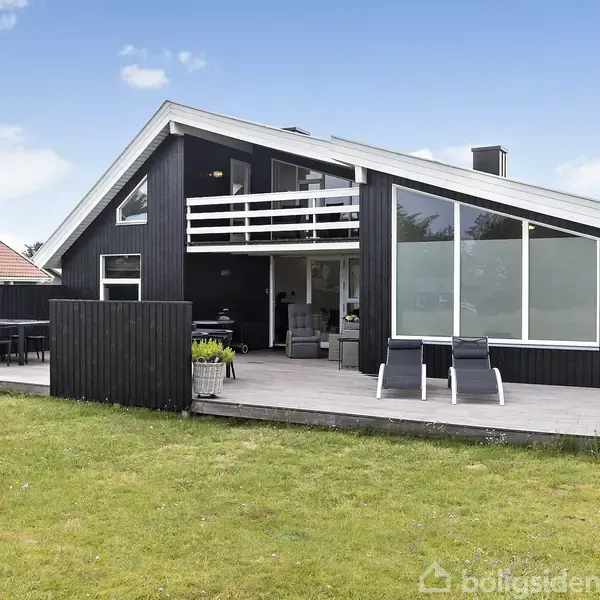 Sort træhus med stor terrasse omgivet af græsplæne. Huset har store vinduer og balkon samt to liggestole på terrassen. Himlen er blå med få skyer.